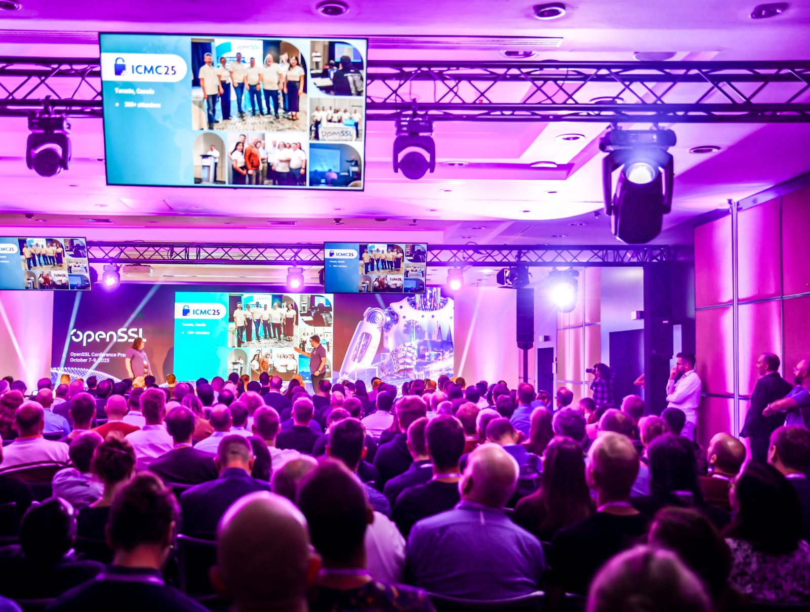 The main hall of the OpenSSL Conference Prague 2025 opening — a packed audience watching the stage as the welcome slides play across overhead screens, lit in conference purple.