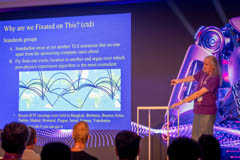 A speaker at the main stage gesturing at a slide reading "Why are we Fixated on This?"